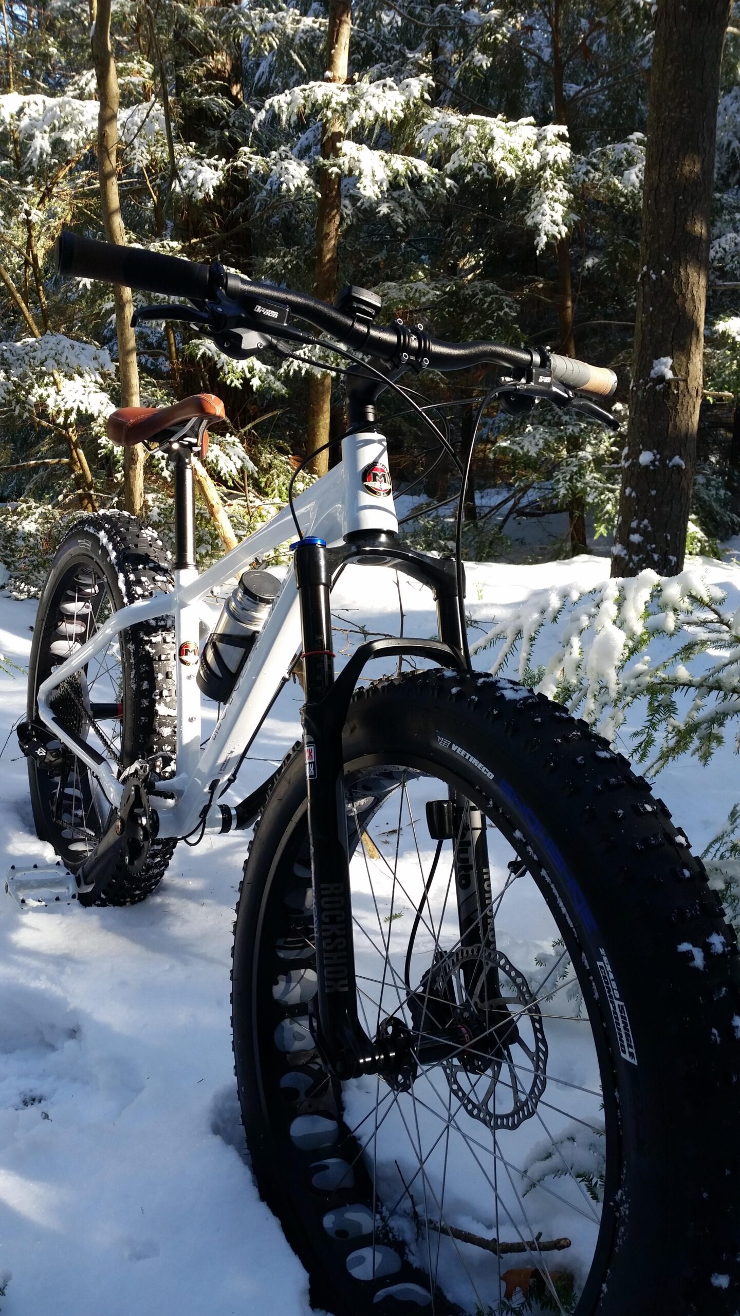 Motobecane Sturgis Bullet: A white fat bike with a brown leather seat parked on snow-covered ground, surrounded by snow-dusted trees. The bike features wide, knobby tires designed for winter riding, and a water bottle mounted on the frame. Sunlight filters through the trees, illuminating the scene.
