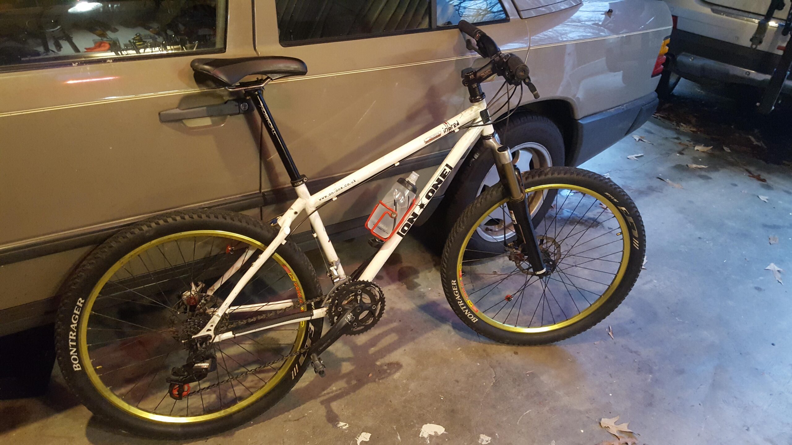 On-One On one inbred: A white mountain bike with yellow accents is leaning against a parked car in a garage setting. The bike features wider tires and a water bottle holder attached to the frame, highlighting its outdoor and recreational use. The garage floor shows some leaves and tools in the background, indicating it's a working space.
