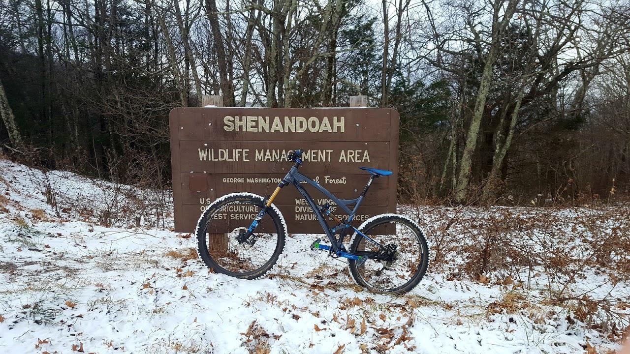 A mountain bike leaning against a wooden sign that reads "Shenandoah Wildlife Management Area" in a snowy landscape with bare trees in the background. Reddish Knob mountain bike trail.