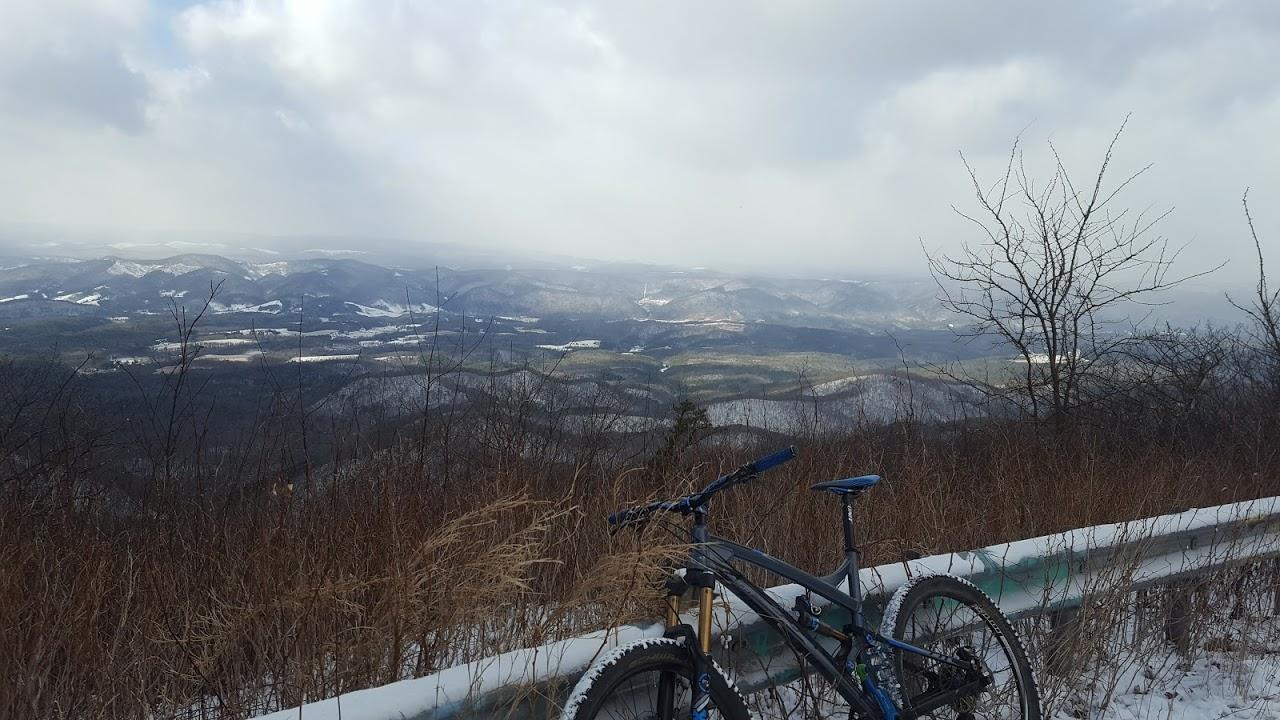 A mountain bike leaning against a guardrail overlooking a scenic view of snow-covered hills and valleys under a cloudy sky. Reddish Knob mountain bike trail.