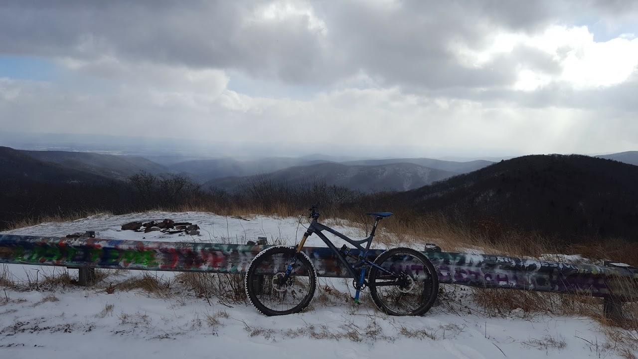 A mountain bike resting on a snow-covered ledge, with a scenic view of rolling hills and valleys in the background. The foreground features a graffiti-covered guardrail, while the sky above is partly cloudy, hinting at an overcast winter day. Reddish Knob mountain bike trail.