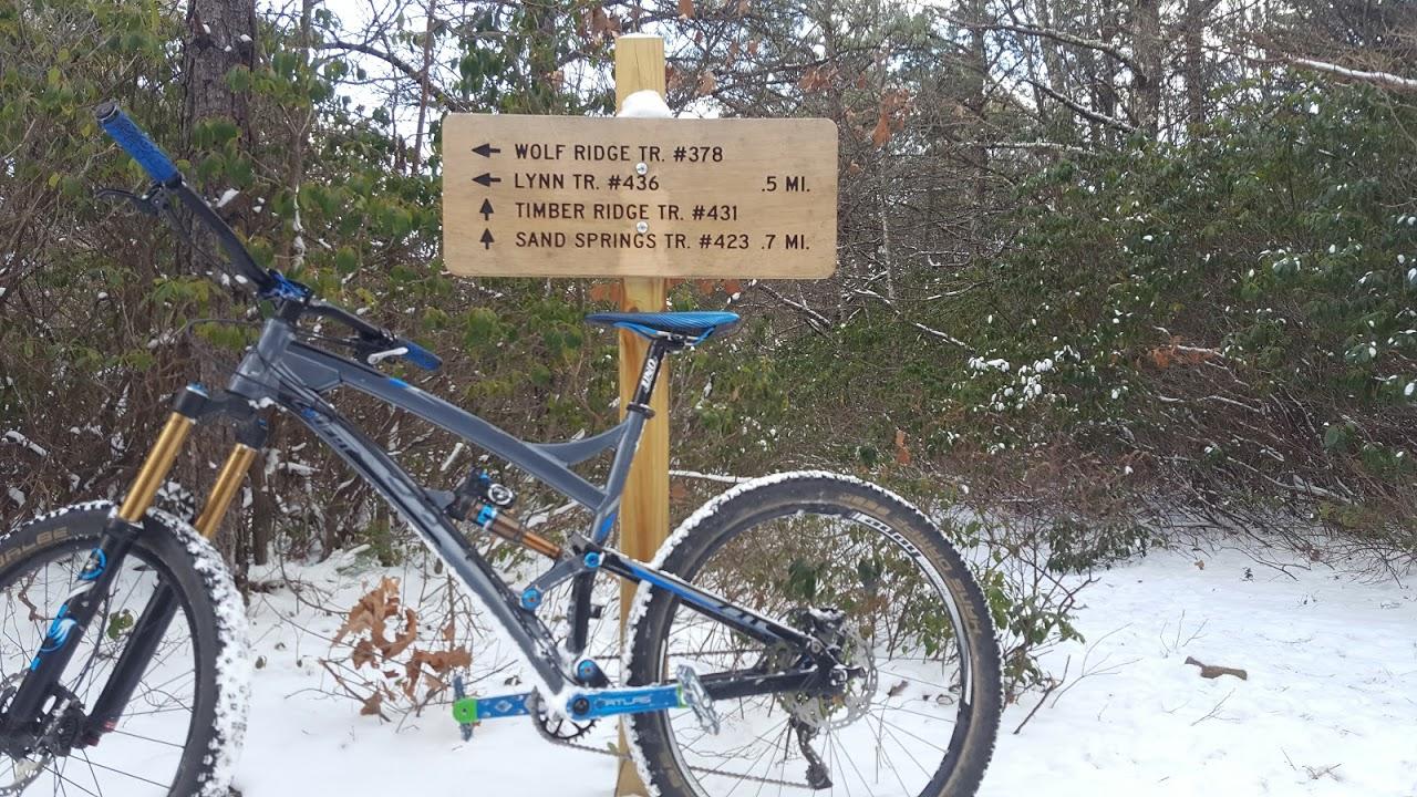 A mountain bike rests on snow-covered ground next to a wooden trail sign. The sign indicates directions and distances for several trails: Wolf Ridge Trail (#378) to the left, Lynn Trail (#436) 0.5 miles, Timber Ridge Trail (#431) straight ahead, and Sand Springs Trail (#423) 0.7 miles. Surrounding the area are trees and brush, creating a scenic winter setting. Reddish Knob mountain bike trail.