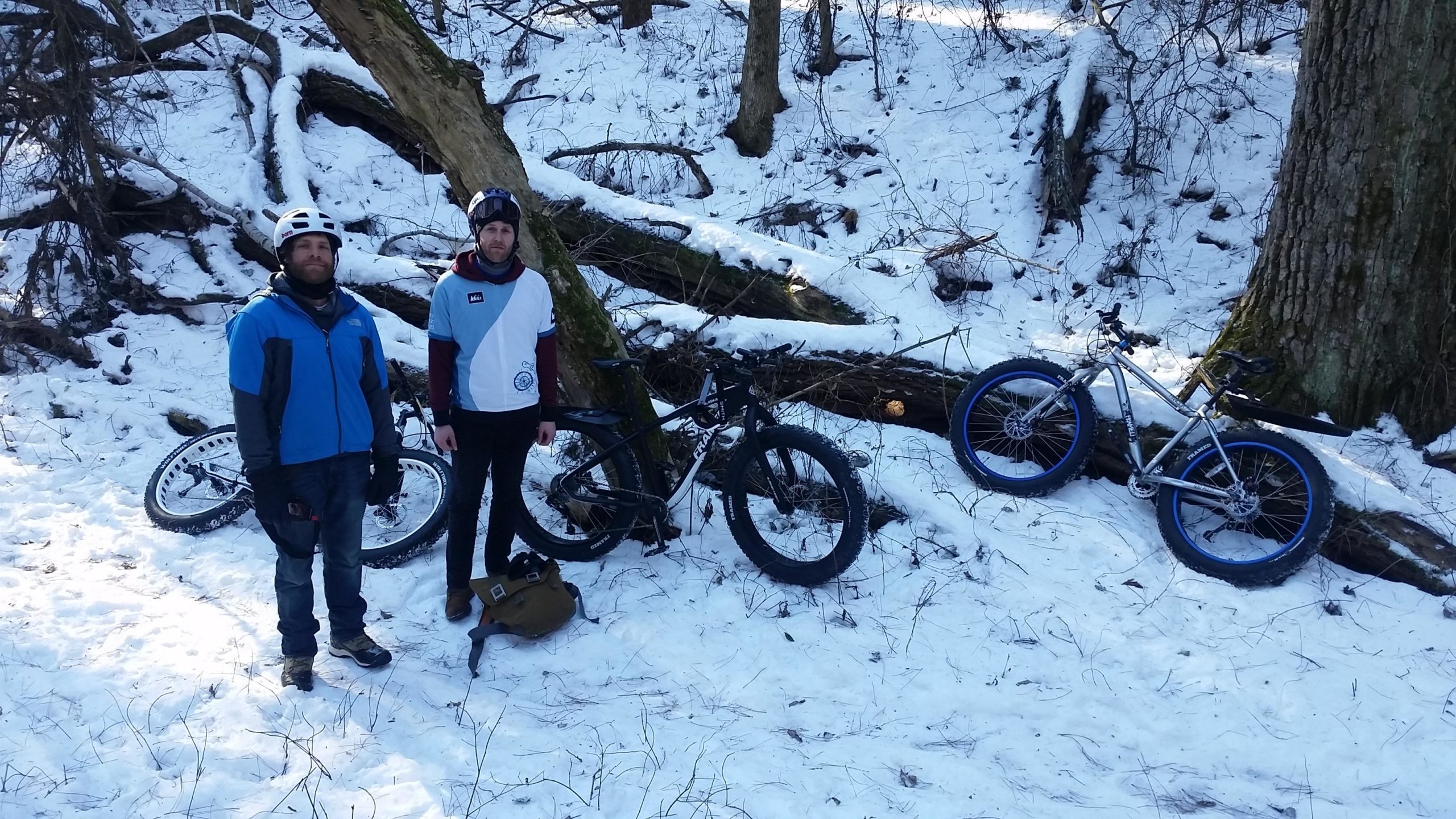 Two mountain bikers stand on a snowy trail surrounded by trees. They are dressed in winter cycling gear, with one wearing a blue jacket and the other in a light blue and maroon shirt. Several bicycles, including a fat bike, are parked nearby in the snow. Reservoir Woods mountain bike trail.