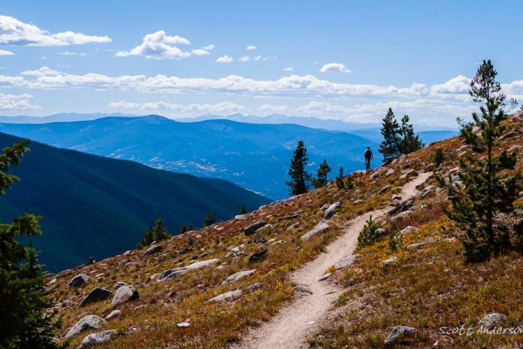 A hiker walks along a winding dirt trail through a mountainous landscape, surrounded by green trees and rocky terrain. The backdrop features a vast view of rolling hills under a bright blue sky with fluffy white clouds. The scene captures the beauty of nature and the serenity of outdoor exploration.