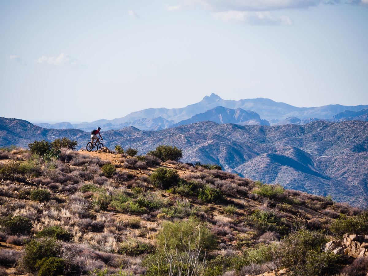 A mountain biker navigating a rocky trail on a hillside, surrounded by rolling mountains and a clear blue sky. The landscape features sparse vegetation, including bushes and cacti, highlighting the rugged terrain of the area. Arizona Trail mountain bike trail.