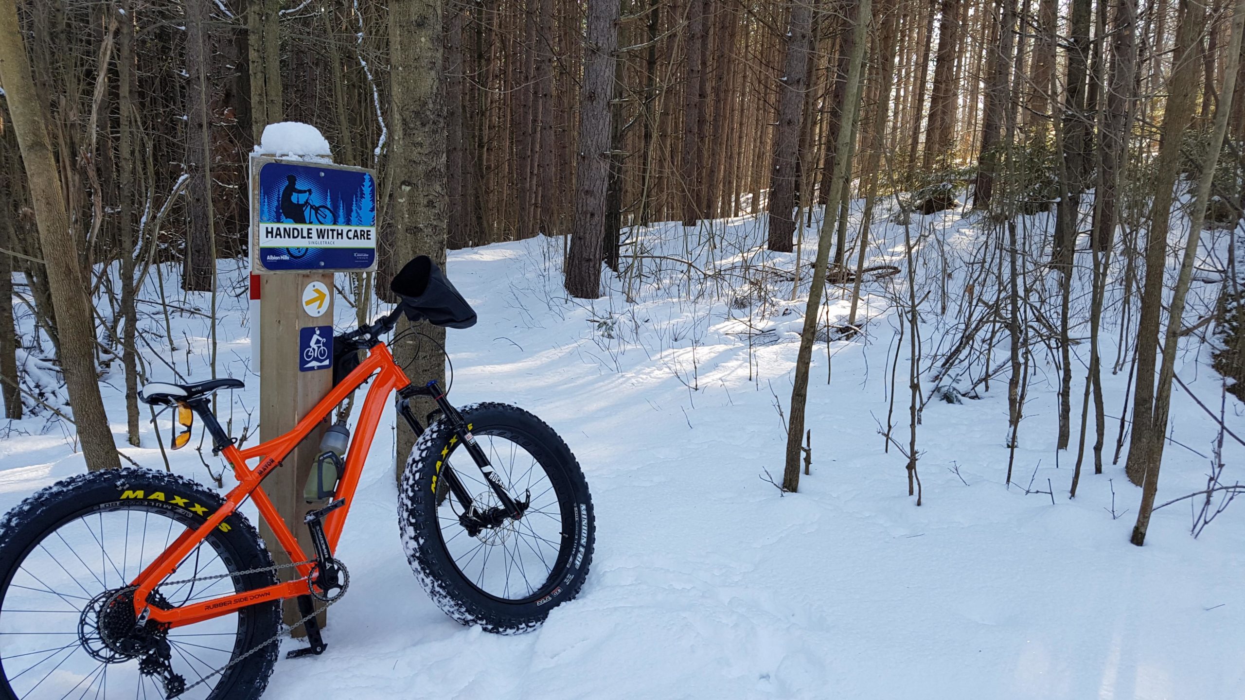 A bright orange fat bike parked next to a snow-covered trail sign that reads "HANDLE WITH CARE." The scene is set in a winter forest with tall trees and a blanket of fresh snow on the ground. The bike features wide tires designed for snowy conditions, and the background shows a peaceful, wintry landscape. Albion Hills mountain bike trail.
