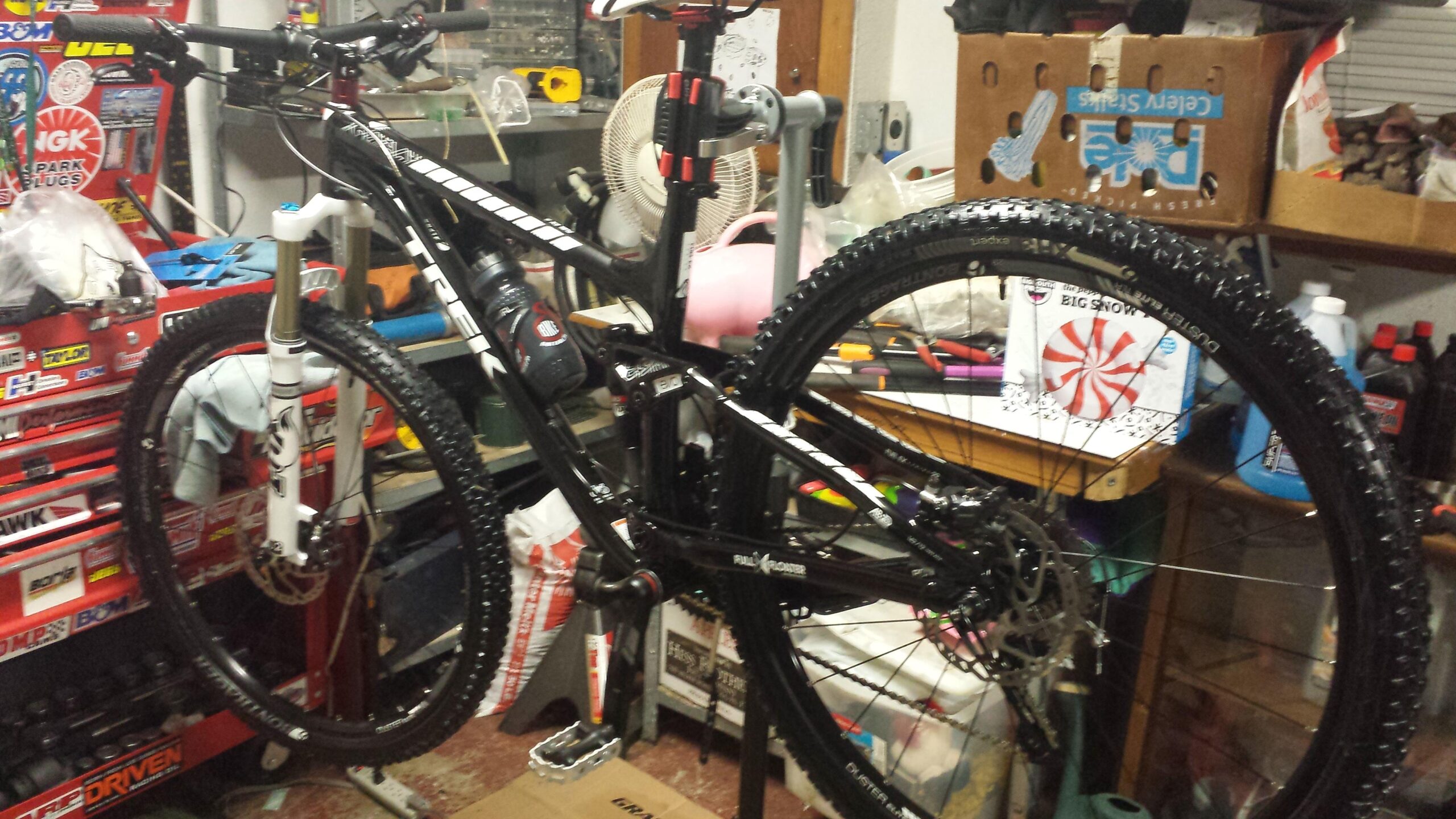 Trek Fuel EX 8: A black mountain bike with a front suspension fork mounted on a repair stand in a cluttered garage workspace. Various tools, parts, and containers are visible in the background, indicating a biking maintenance or assembly area. The bike features thick, textured tires and a water bottle attached to the frame.