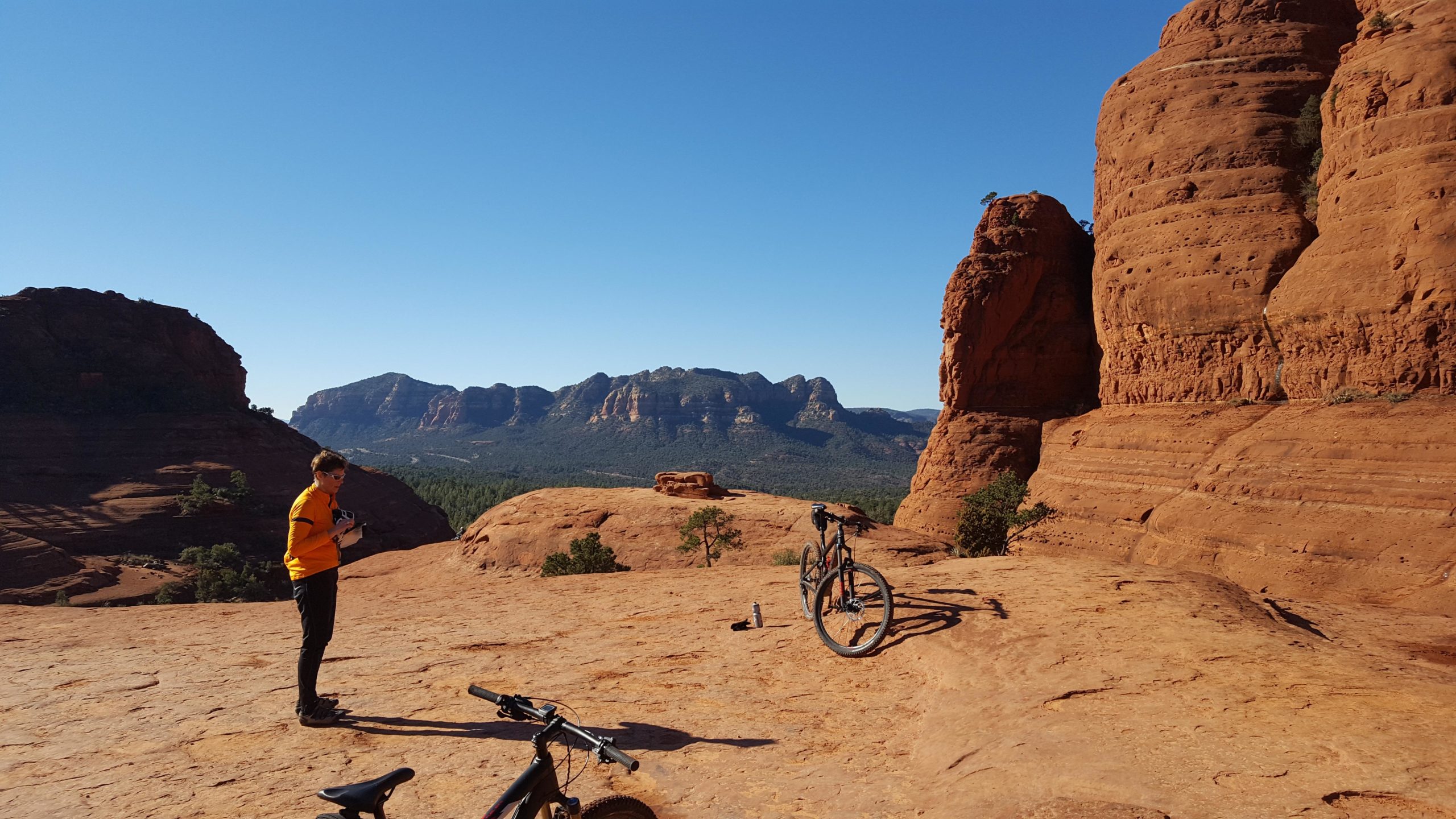 A person in an orange shirt stands on a rocky landscape with red sandstone formations in the background. Two mountain bikes are positioned nearby, and a scenic view of distant mountains and a clear blue sky are visible. Broken Arrow Trail / Chicken Point mountain bike trail.