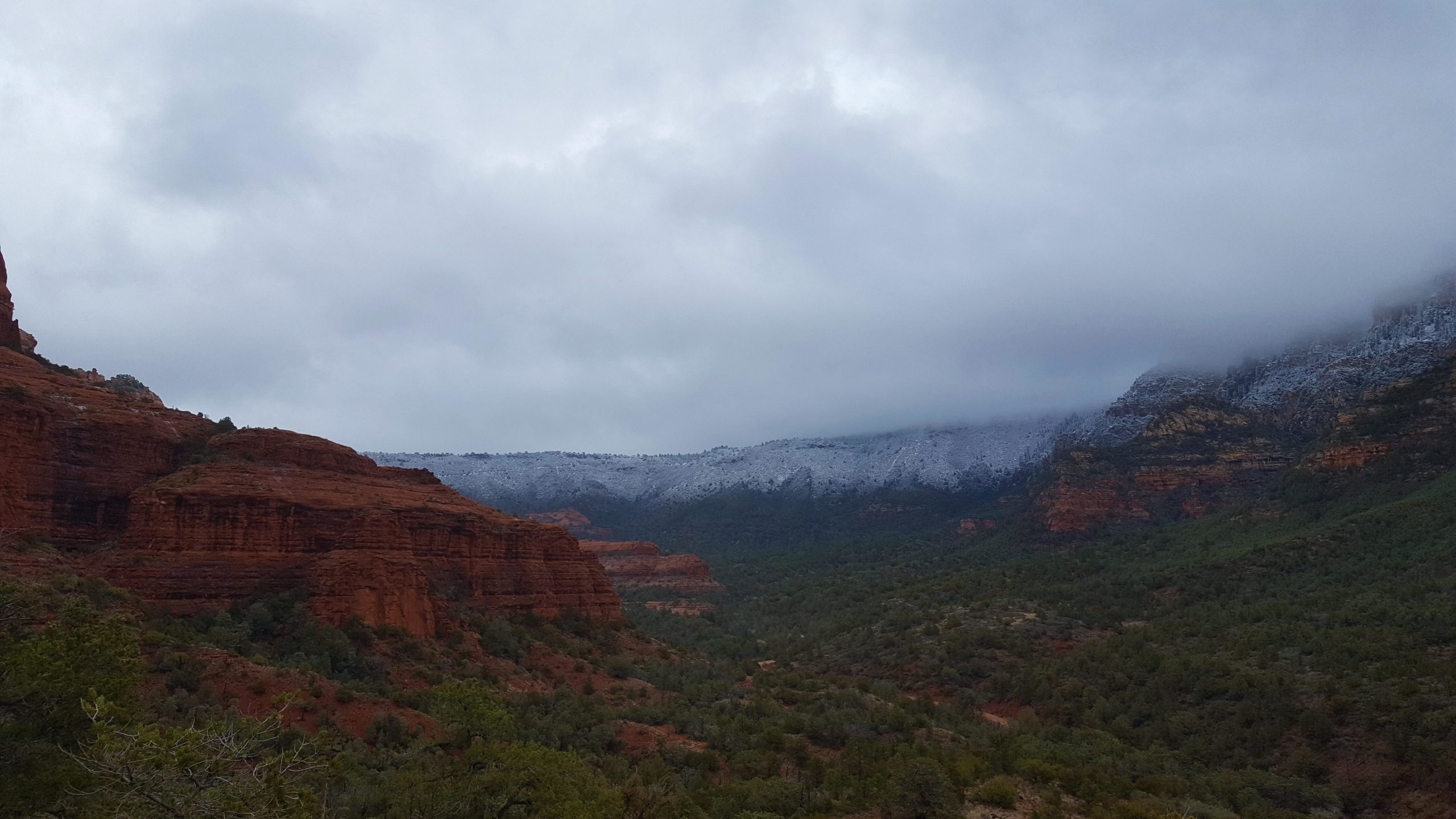 A panoramic view of a rugged landscape featuring red rock formations and green vegetation, with a layer of snow covering the mountains in the background under a cloudy sky. Hangover mountain bike trail.