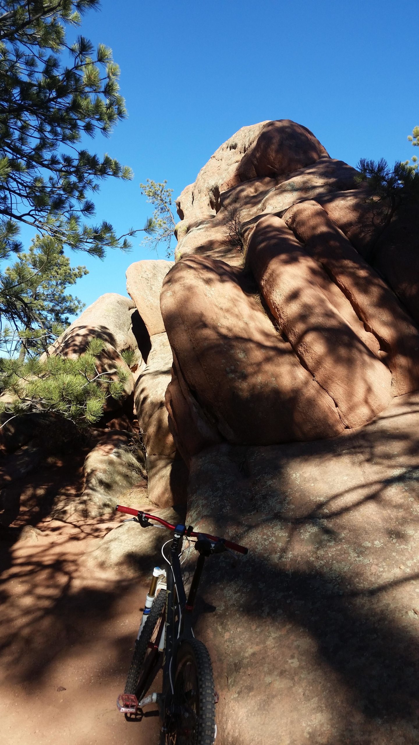 A mountain bike leaning against large rocks in a natural landscape under a clear blue sky. Pine tree branches are visible in the upper left corner, casting shadows on the rocky surface. The scene captures an adventurous outdoor setting suitable for biking or hiking. Red Rock Canyon mountain bike trail.