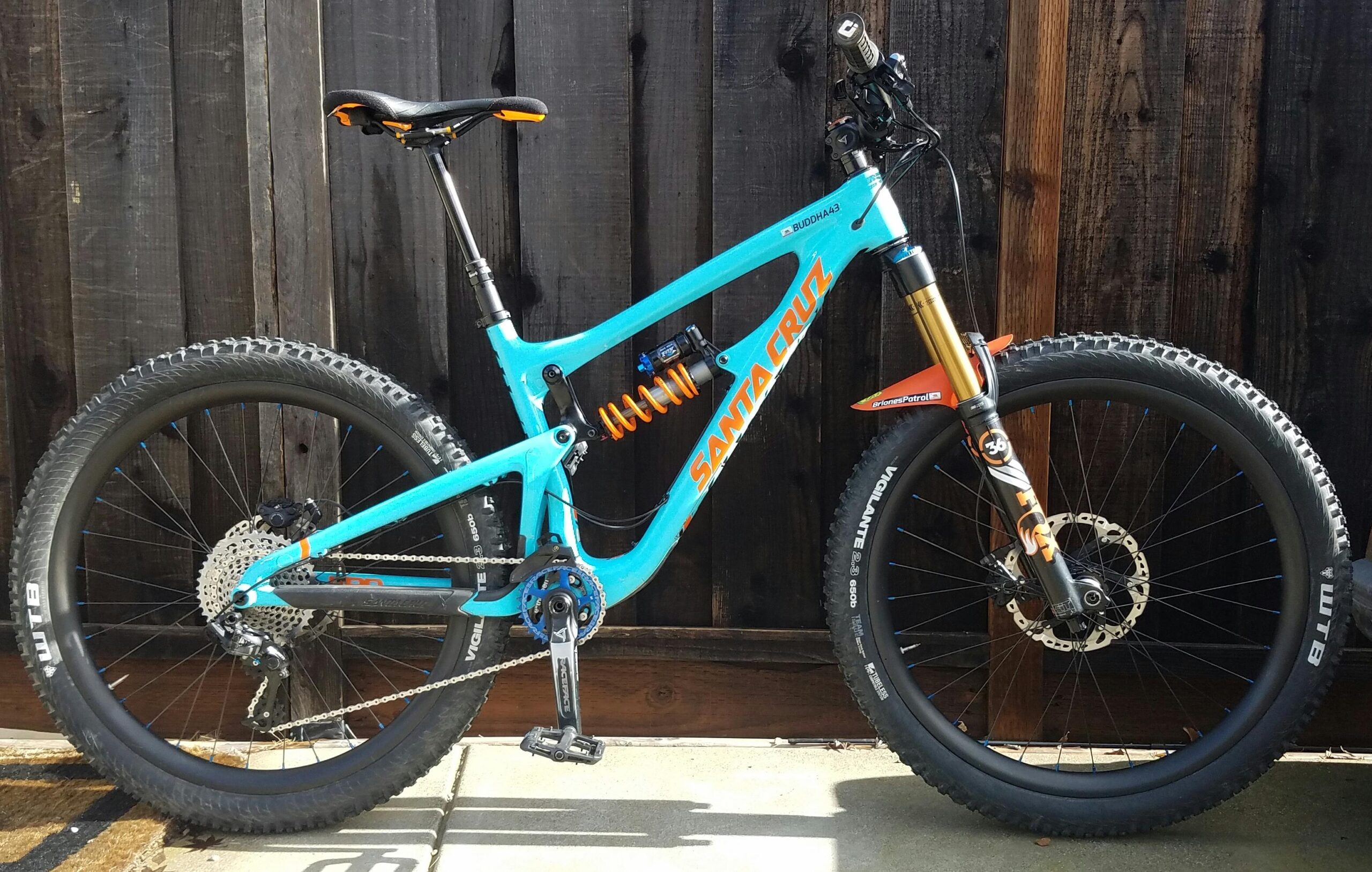 Santa Cruz Nomad Carbon 27.5: A bright blue Santa Cruz mountain bike with a sleek design, featuring black wheels, an adjustable seat, and an orange rear shock. The bike is positioned against a wooden background, showcasing its detailed components such as the drivetrain and suspension system.