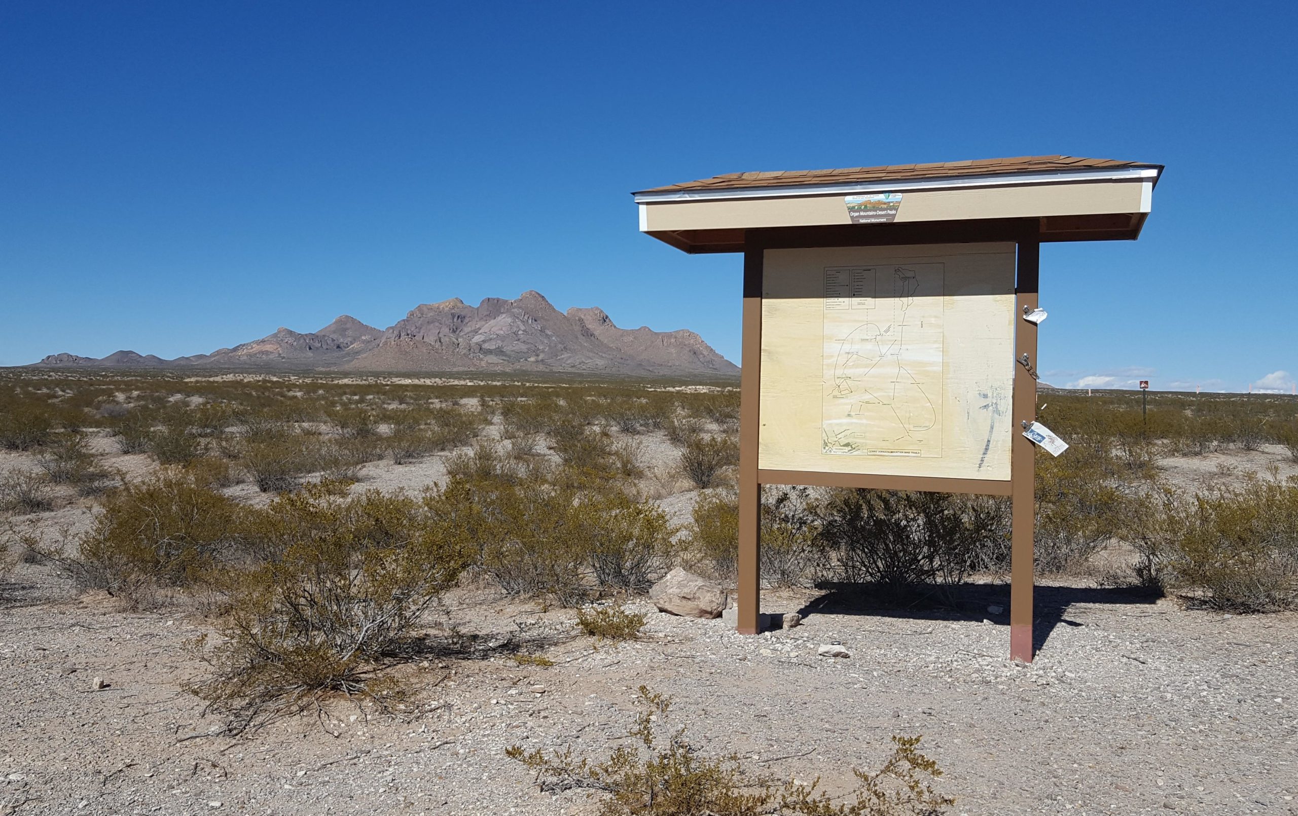 A informational signpost stands in a desert landscape with sparse vegetation, featuring a map on its surface. In the background, rugged mountains rise under a clear blue sky. The scene conveys a sense of remote natural beauty and exploration. Dona Ana mountain bike trail.