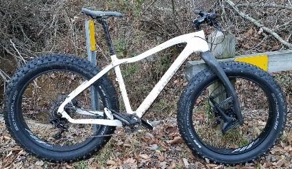 Framed Alaskan Carbon: A white fat bike with oversized tires parked next to a wooden post in a wooded area. Fallen leaves cover the ground, and there are hints of trees and brush in the background.