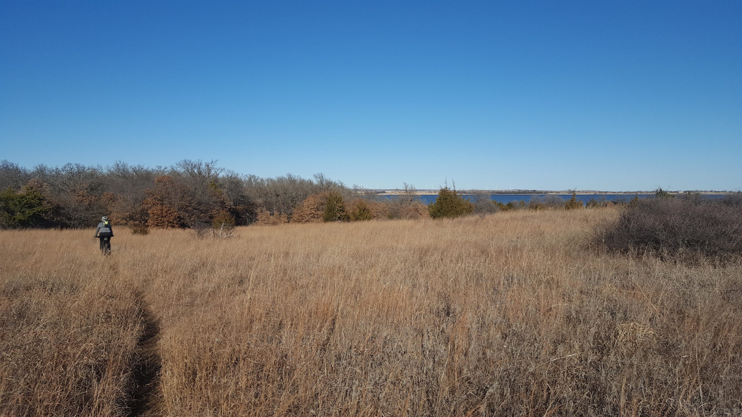 A person walking along a dirt path through tall, dry grass in a natural landscape, with trees and a body of water visible in the background under a clear blue sky. Lake Lawtonka Trails mountain bike trail.