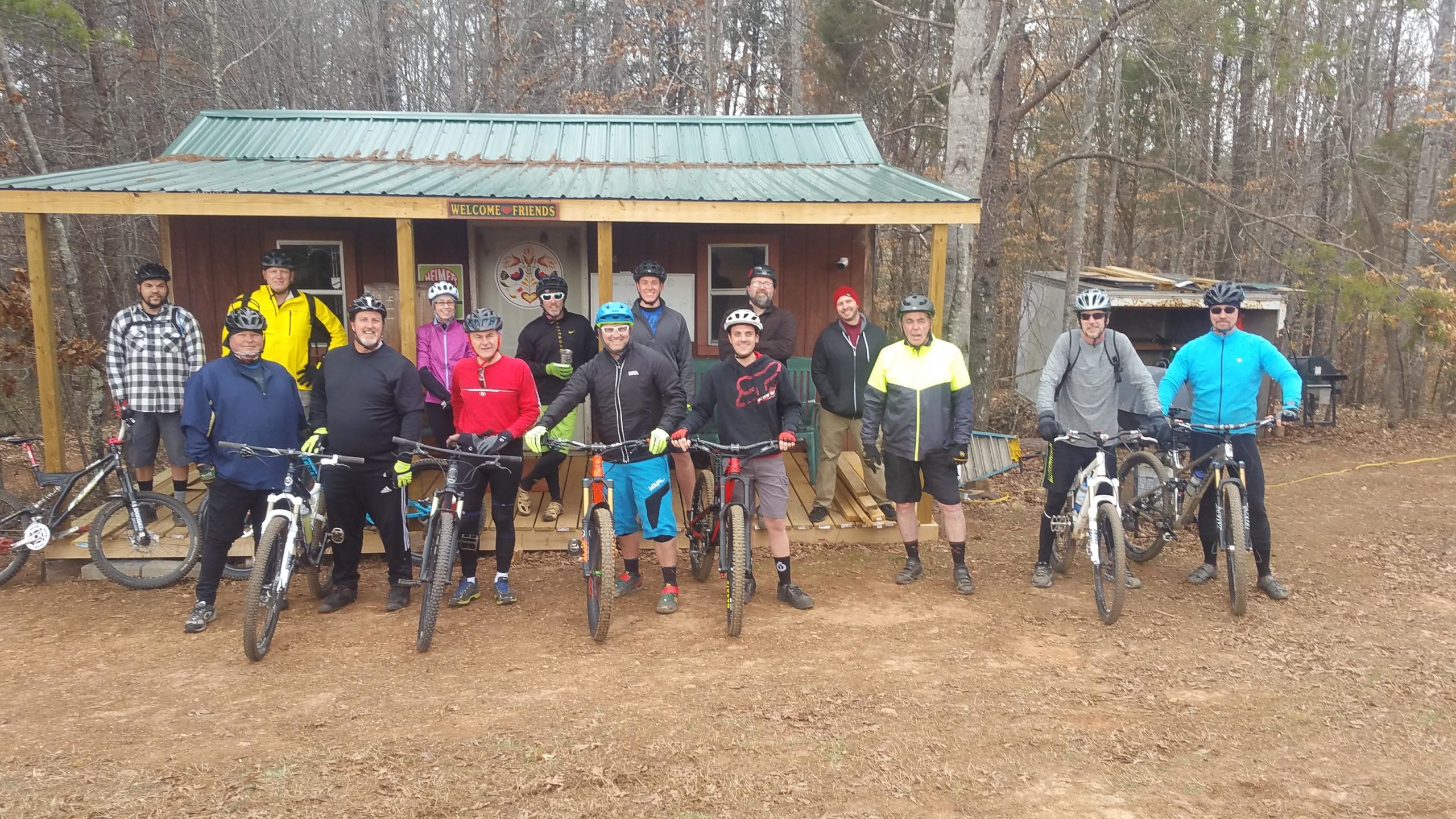 A group of eleven mountain bikers posing with their bikes in front of a wooden shelter with a green metal roof. The group is wearing a mix of cycling gear, including helmets and bright jackets, and is set in a forested area with bare trees in the background. The mood is friendly and casual. Mountain Laurel Trails mountain bike trail.