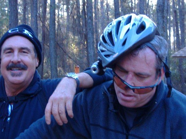 Specialized SWORKS Stumpjumper FSR: Two men are standing outdoors in a wooded area. One man is smiling and has his arm around the shoulder of the other, who appears to be adjusting a bike helmet that is slightly askew on his head. The second man is wearing sunglasses and looks amused. The setting is bright, with trees visible in the background, indicating an active outdoor environment.