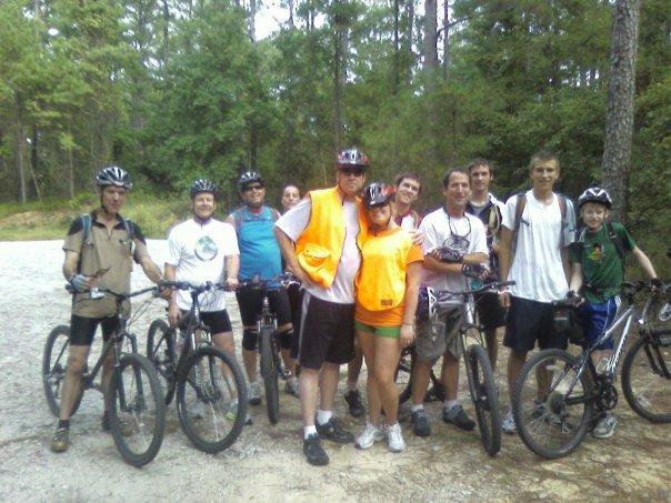 Specialized SWORKS Stumpjumper FSR: A group of ten people stands together on a dirt path surrounded by trees, all wearing helmets and preparing for a mountain biking adventure. They are posing with their bicycles, and a few individuals are wearing brightly colored safety vests. The group appears cheerful and ready for outdoor activities.