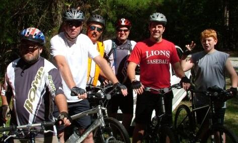 Specialized SWORKS Stumpjumper FSR: A group of six men stands together with their mountain bikes in a wooded area, wearing helmets and casual cycling attire. They appear to be taking a break during a biking outing, with one man playfully gesturing with his peace sign. The scene is bright and sunny, showcasing a casual outdoor atmosphere.
