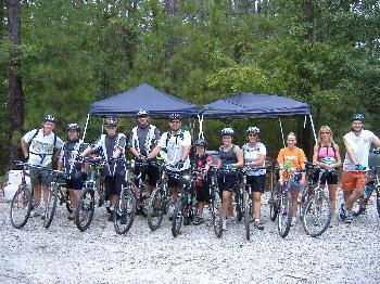 Specialized SWORKS Stumpjumper FSR: A group of ten individuals, all wearing helmets, stand beside their bicycles in a forested area. Two black canopies are set up in the background. The group is dressed in casual cycling attire, smiling and posing for the photo, with trees surrounding them.