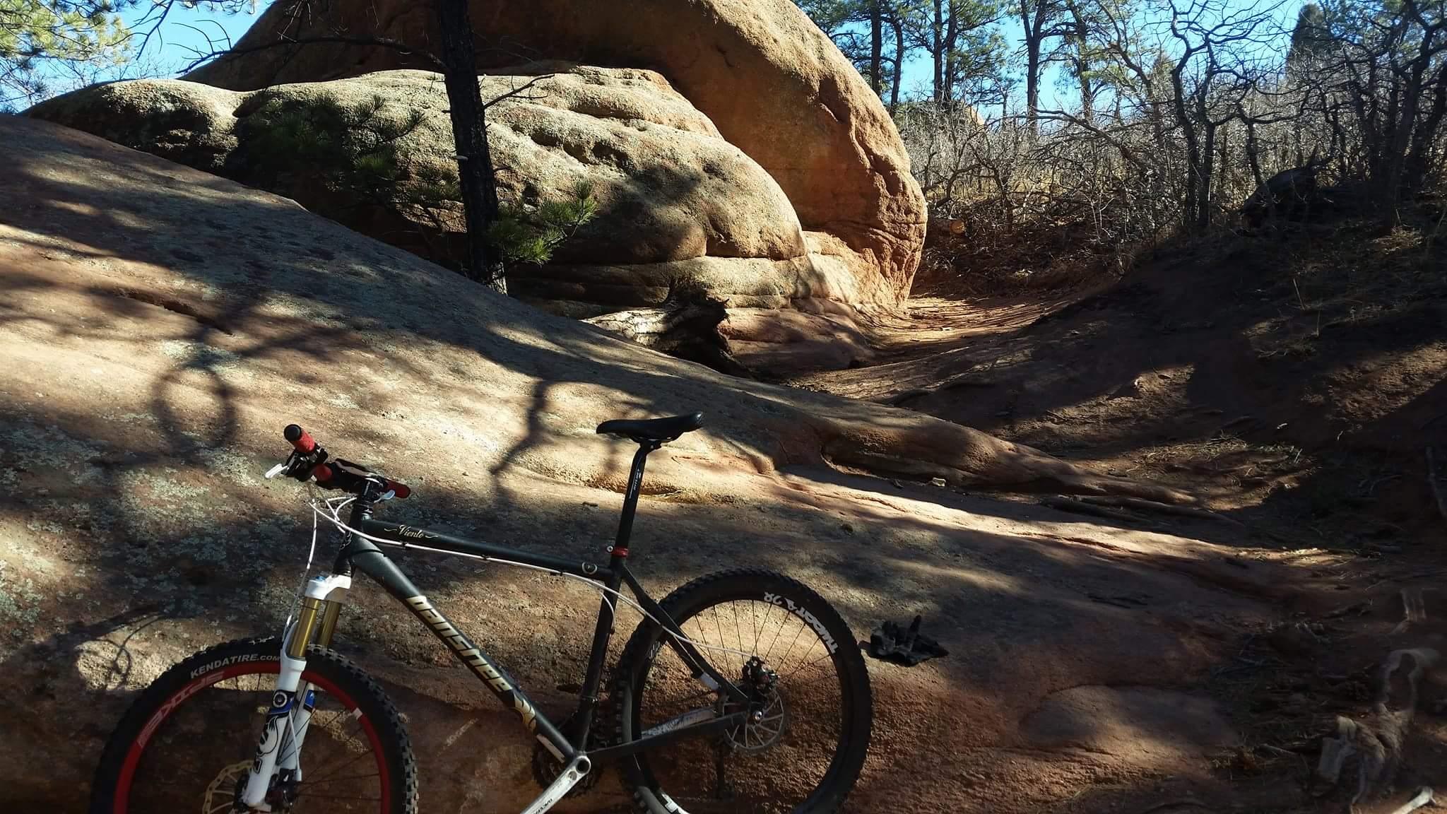 A mountain bike resting on rocky terrain, surrounded by trees and large boulders. The scene captures a trail with a mixture of natural features, including smooth rock surfaces and scattered vegetation, under a clear blue sky. Red Rock Canyon mountain bike trail.