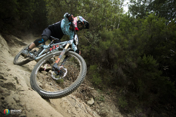 A mountain biker expertly navigating a sandy trail, leaning into a turn with intense focus. The rider is wearing protective gear, including a helmet and gloves, and appears to be racing through a wooded area with vibrant green foliage surrounding the path. The bike features a sturdy frame and thick tires, highlighting the rugged terrain.