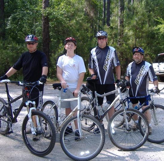 Specialized SWORKS Stumpjumper FSR: Four cyclists pose with their mountain bikes in a forested area. They are wearing helmets and cycling attire, with two men in black shirts, one in a white shirt, and another in a black and white cycling jersey. The background features green trees, indicating a natural outdoor setting.