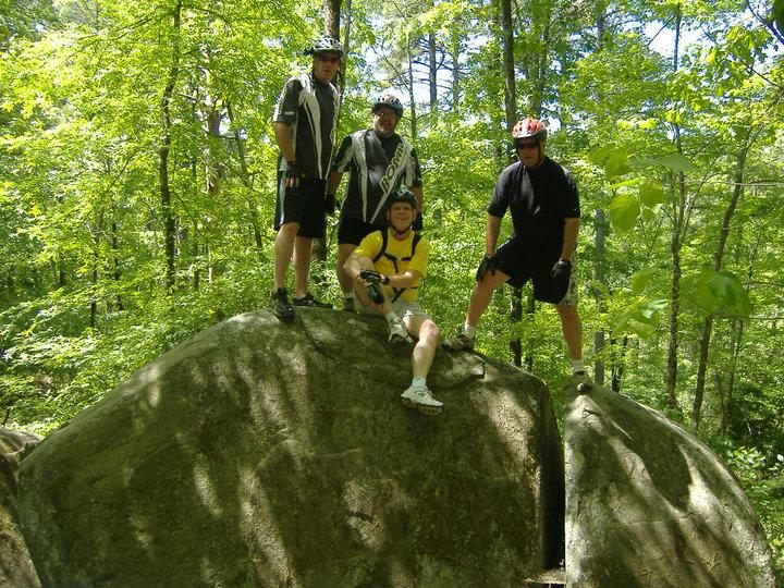 Specialized SWORKS Stumpjumper FSR: Four mountain bikers pose on a large rock in a forested area. They are wearing helmets and biking gear, with trees and greenery surrounding them. One person is seated on the rock, while the others stand around him, all smiling and enjoying the outdoors.