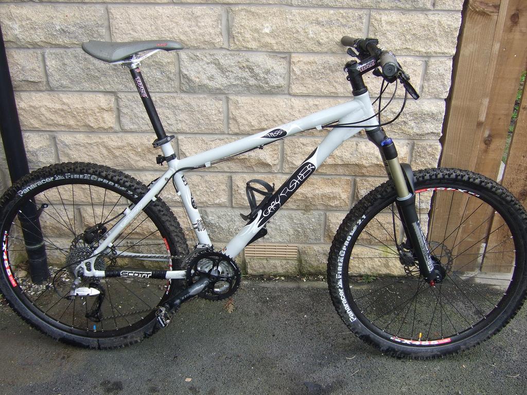 Gary Fisher Tassajara: A gray mountain bike with a black frame, featuring thick tires and suspension forks, is leaned against a stone wall. The bike has a textured seat, hand brakes, and gear shifters, appearing well-maintained and ready for off-road riding.