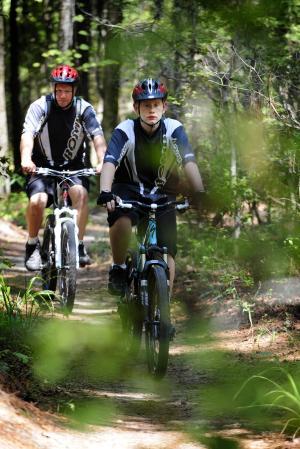 Specialized SWORKS Stumpjumper FSR: Two cyclists riding mountain bikes on a wooded trail, surrounded by greenery. One cyclist is ahead, while the other follows closely behind. Both are wearing helmets and cycling jerseys.
