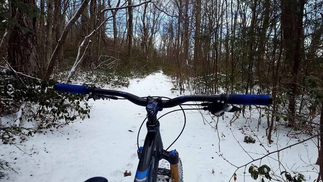 A view of mountain bike handlebars positioned in the foreground, with a snow-covered trail ahead and surrounded by trees and underbrush in a winter forest setting. Reddish Knob mountain bike trail.