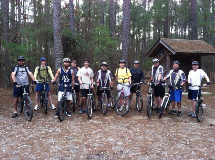 Specialized SWORKS Stumpjumper FSR: Group of ten people wearing helmets, standing with mountain bikes on a dirt path in a forested area. They are posing together, with a small wooden structure visible in the background. The scene conveys a sense of camaraderie and outdoor adventure.