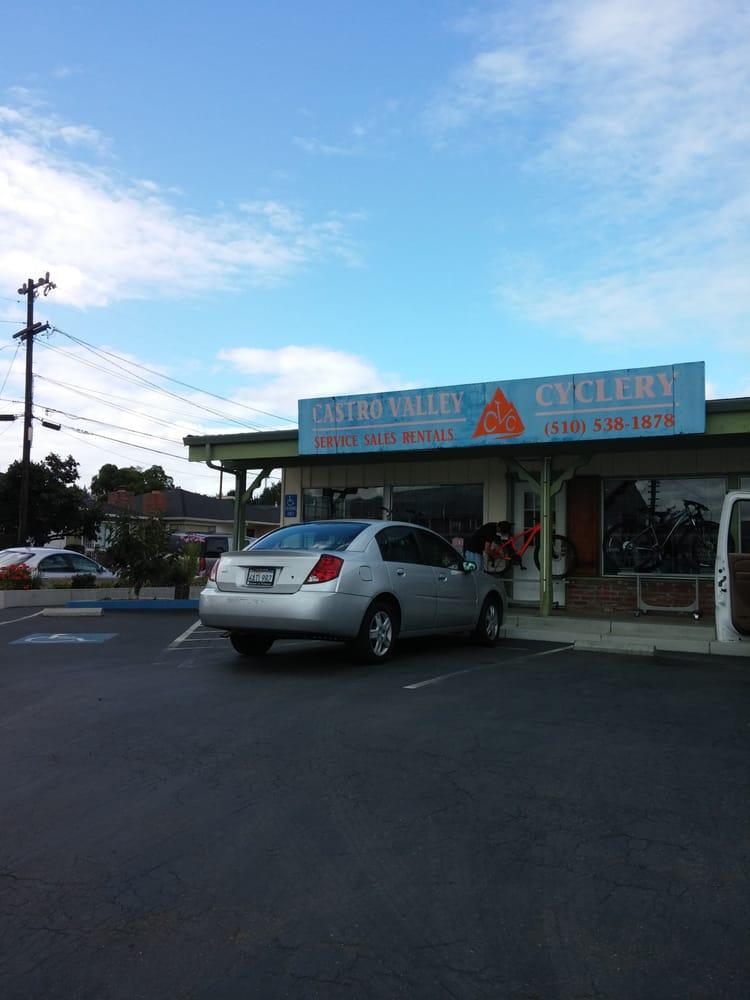 A storefront of Castro Valley Cyclery, featuring a blue sign that reads "CASTRO VALLEY CYCLERY - SERVICE SALES RENTALS" along with a contact number. In the foreground, a silver car is parked near the entrance, and a person is seen engaging with a bike outside the shop. The background features a clear blue sky with some clouds.