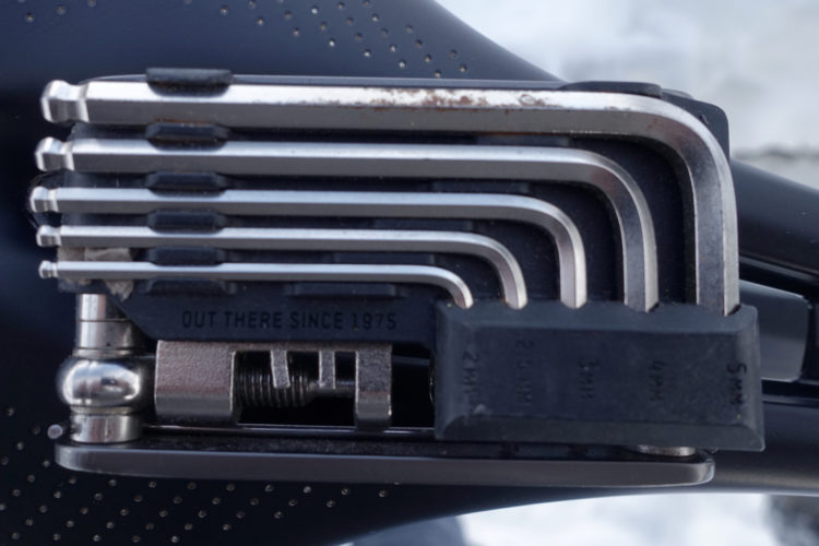 A close-up image of a multi-tool featuring several Allen wrenches stacked together, along with a small flat-head screwdriver. The tool is attached to a black background, and the text "OUT THERE SINCE 1975" is visible on the side of the tool.