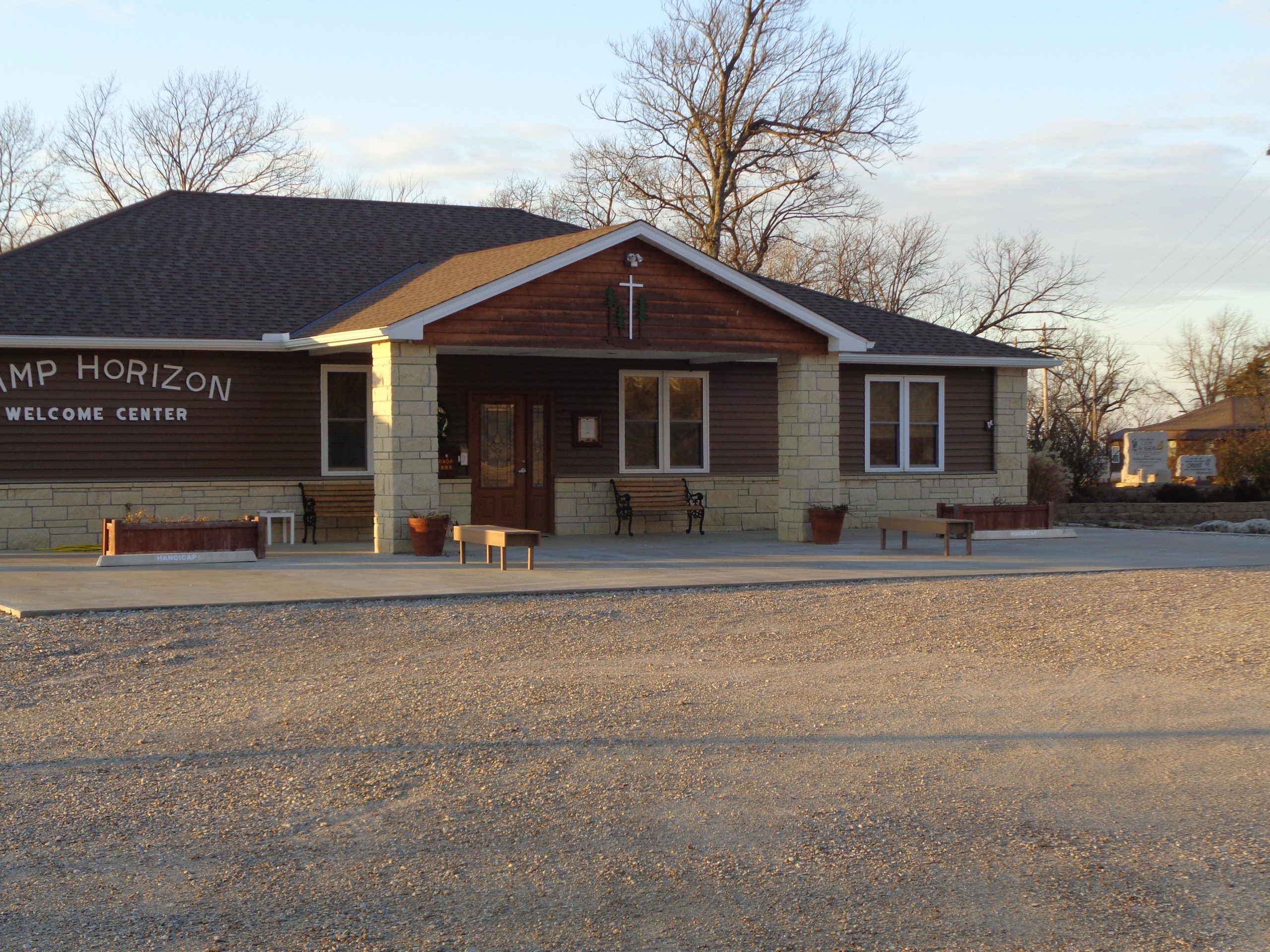 A welcoming center building for Camp Horizon, featuring a mix of wood and stone architecture, surrounded by a gravel parking area. The building has large front windows, benches, and a sign that reads "Camp Horizon Welcome Center." The scene is set against a clear sky with trees in the background. Camp Horizon mountain bike trail.