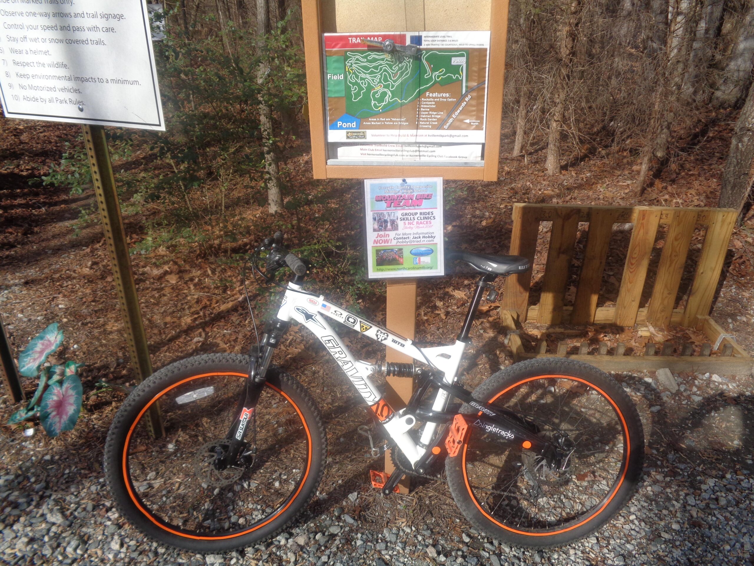 A mountain bike is parked beside a trailhead sign in a wooded area. The sign displays rules and guidelines for trail usage, and there