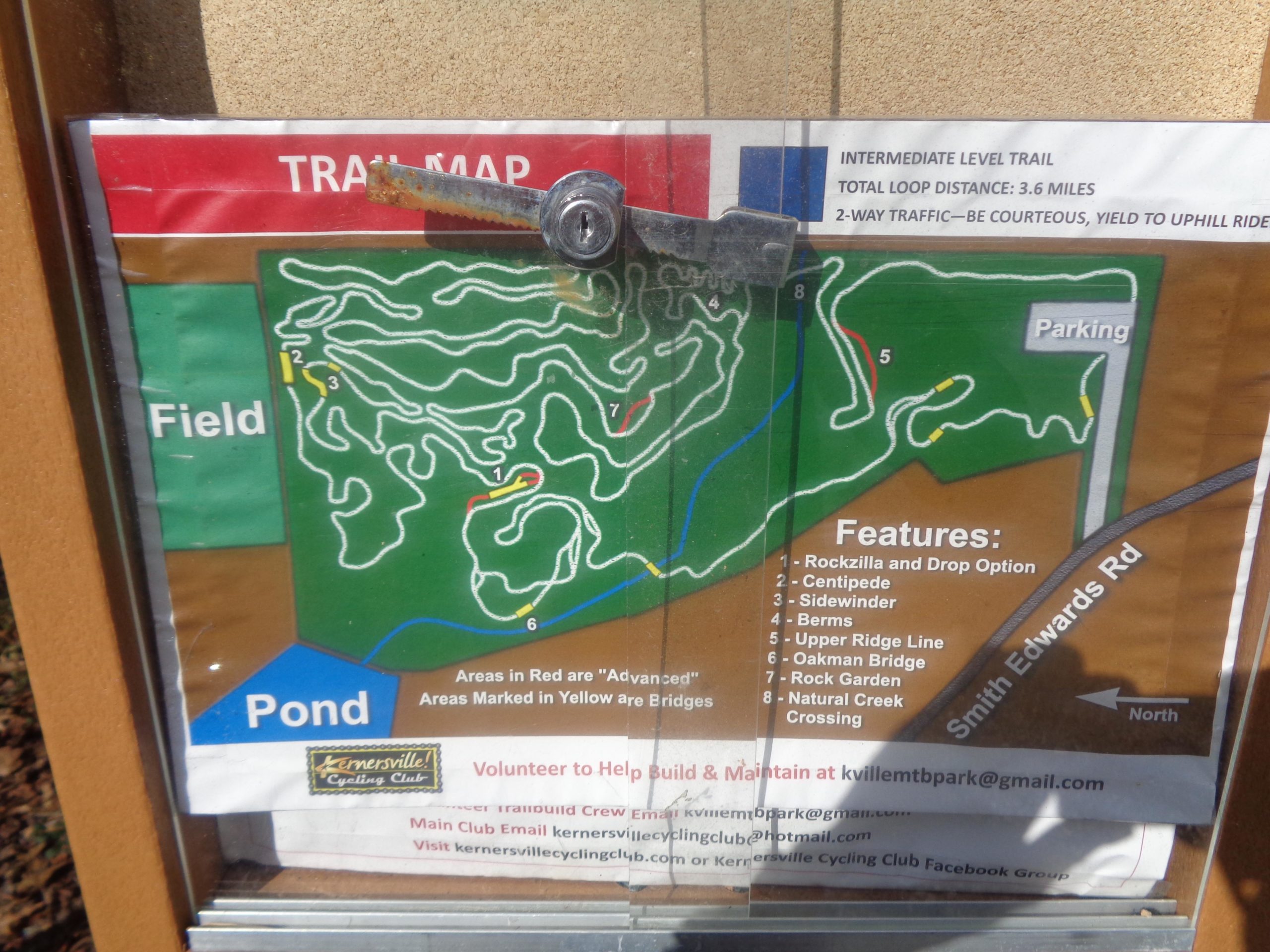 A trail map displayed in a protective case, showing an intermediate-level biking trail with a total loop distance of 3.6 miles. The map outlines various trail features and sections, including areas marked in red for advanced trails and yellow for easier bridges. Key locations such as "Rockzilla," "Centipede," and "Oakman Bridge" are indicated, along with a parking area and nearby road labeled "Smith Edwards Rd." The map is positioned on a wooden post at a park. Kernersville MTB park mountain bike trail.