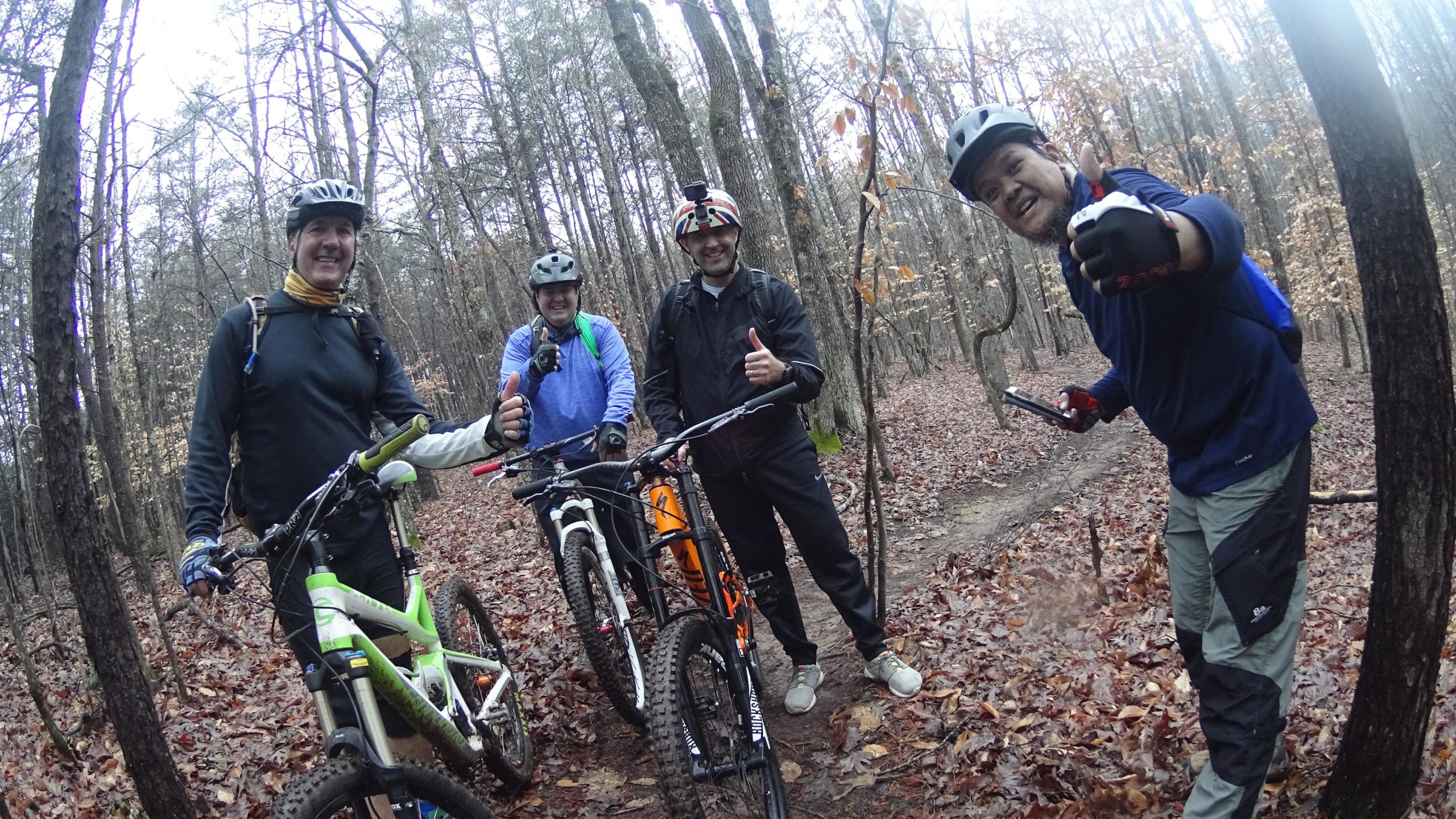 Four cyclists posing with their mountain bikes in a wooded area during a ride, all smiling and giving thumbs up. The scene features tall trees and a forest floor covered with leaves, suggesting an outdoor adventure in nature. Mountain Laurel Trails mountain bike trail.