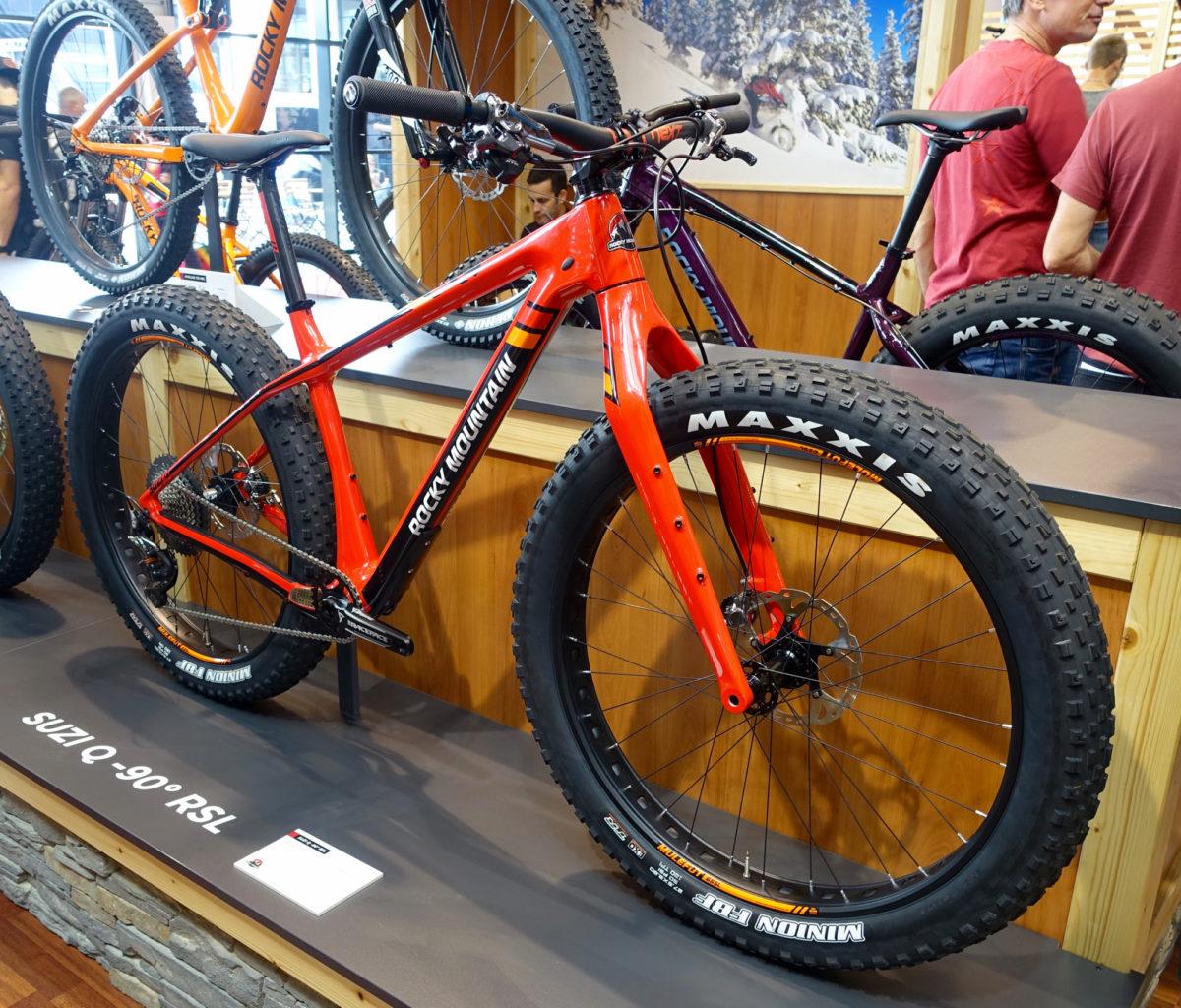 Rocky Mountain Suzi Q: A bright red Rocky Mountain fat bike displayed in a shop, featuring oversized tires for enhanced stability and traction. The bike is positioned on a wooden shelf with a backdrop of winter scenery and other bicycles in the background. The model name "SUZI Q 90° RSL" is visible on the display.