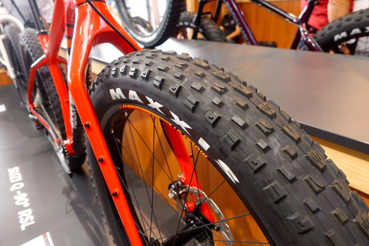 Rocky Mountain Suzi Q: Close-up view of a mountain bike tire on a display, featuring a large, textured tread pattern. The bike frame is primarily orange, showcasing its design. In the background, additional bikes are partially visible along with a wooden display surface.