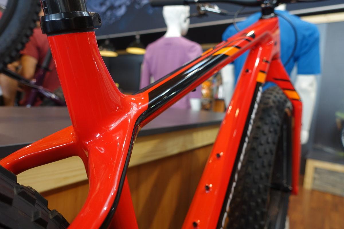 Rocky Mountain Suzi Q: Close-up view of a brightly colored red bicycle frame showcasing sleek design lines and a glossy finish, with a blurred background that includes display mannequins and a wooden counter.