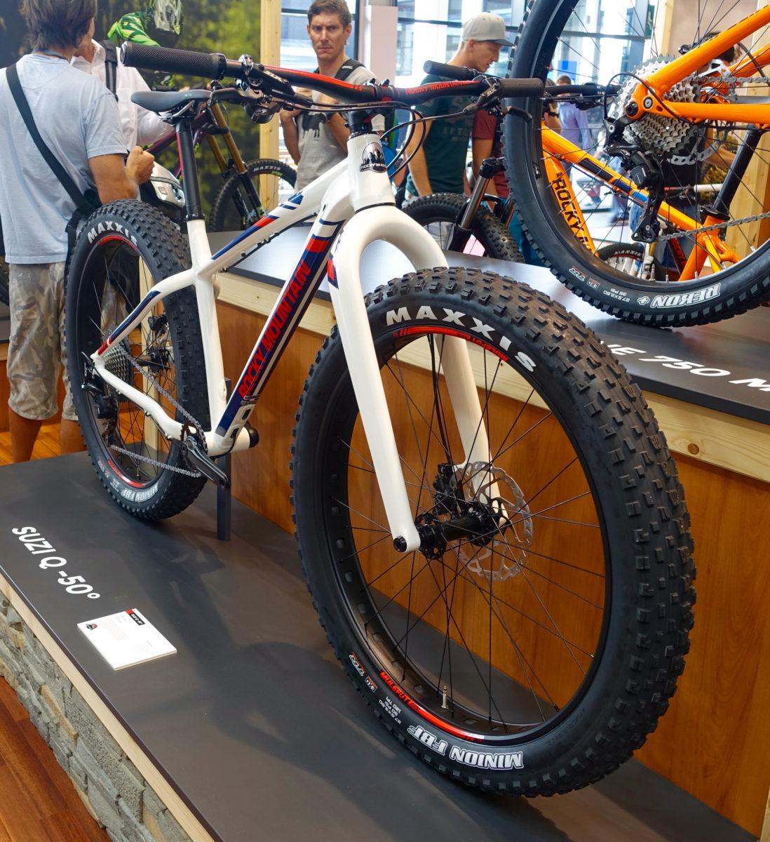 Rocky Mountain Suzi Q: A white mountain bike with wide tires and a prominent design is displayed on a stand in an exhibition area. The bike features a blue and red accent, showcasing the 'Rocky Mountain' brand. In the background, several people observe the bikes on display, with another mountain bike in orange visible.