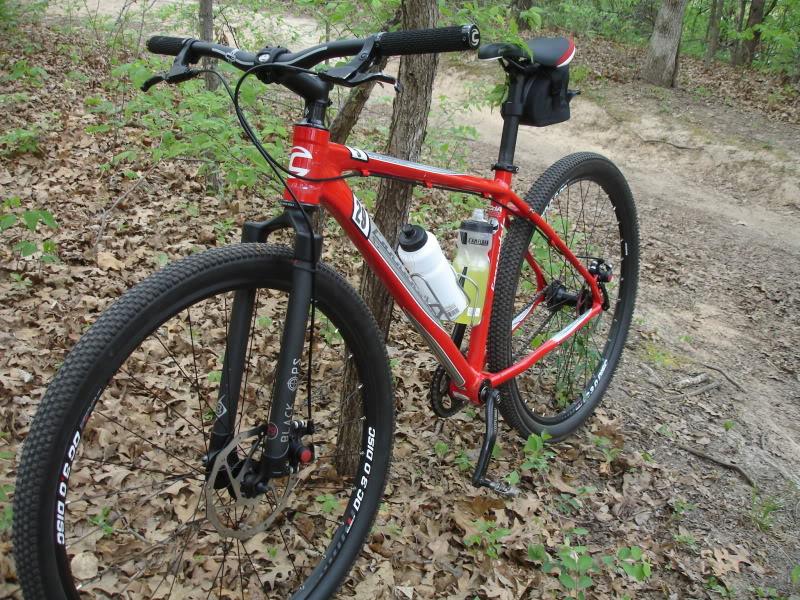 Cannondale Trail 29er 5: A red mountain bike parked against a tree in a wooded area, surrounded by fallen leaves. The bike features sturdy tires, front suspension, and has a water bottle holder attached. A small black saddle bag is mounted on the back. A dirt path is visible in the background, suggesting a trail for cycling.