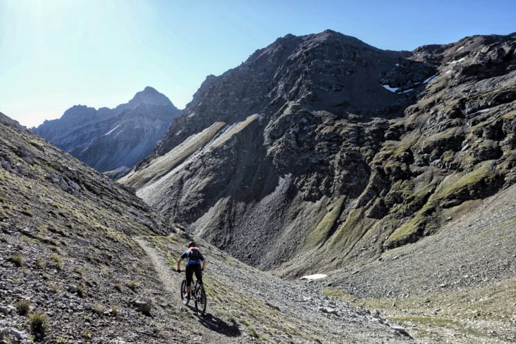 A mountain biker navigates a rugged trail through a scenic mountainous landscape, surrounded by towering peaks and rocky terrain under a clear blue sky.