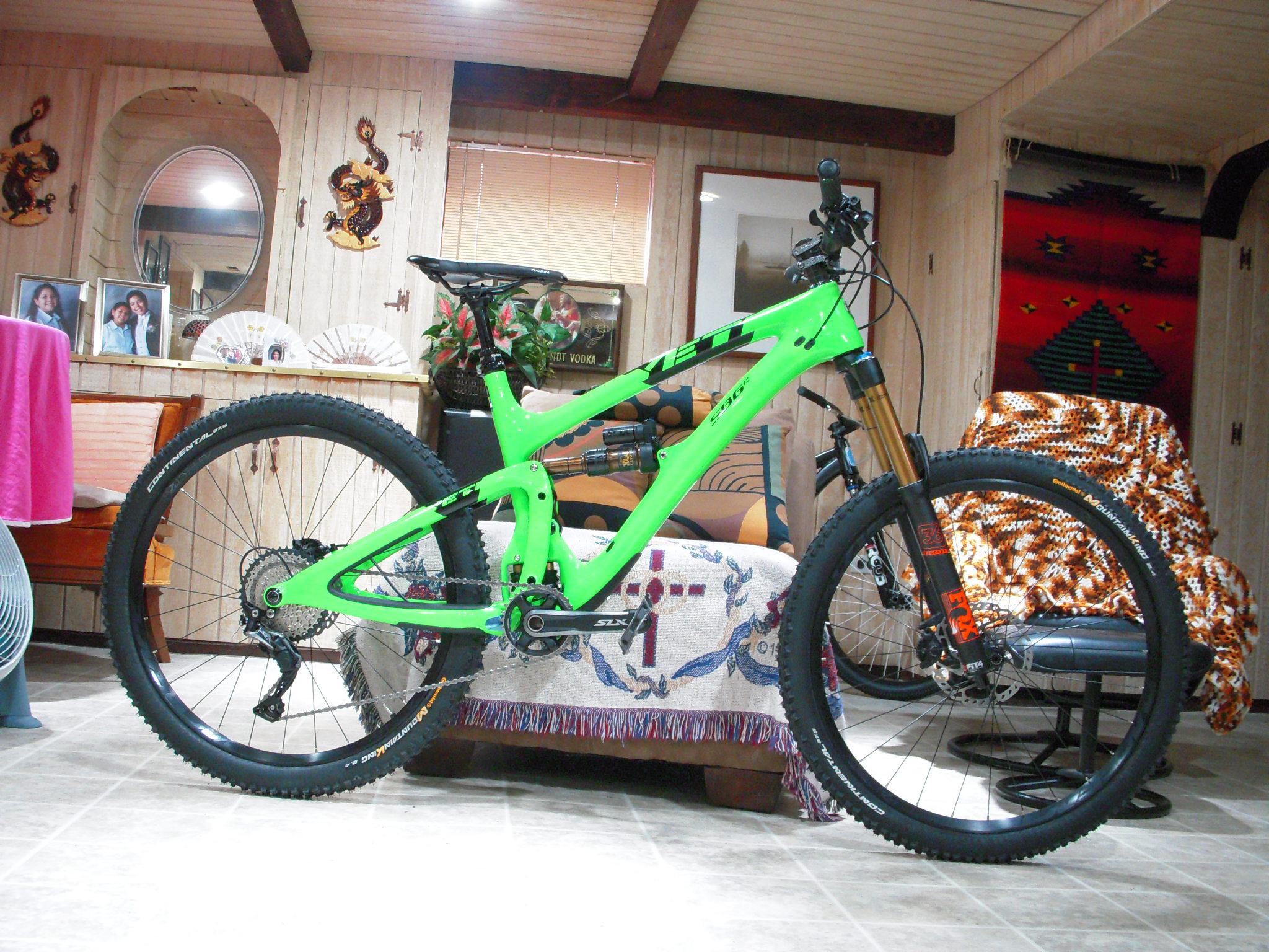 Yeti SB6: A bright green mountain bike with thick tires is positioned indoors against a backdrop of wooden paneling and decorative items, including photos and plants. The bike features a prominent front suspension fork and is placed on a tiled floor, with a couch and a fan visible in the background.