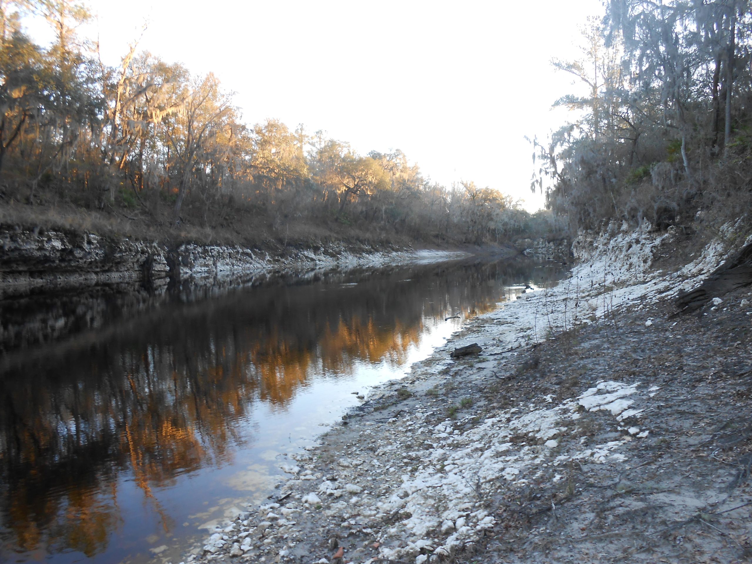A serene view of a riverbank with calm waters reflecting the golden light of sunset. The landscape features rocky banks and trees with sparse leaves, suggesting a tranquil, natural setting. Lush vegetation lines the river, and the scene conveys a sense of peacefulness and solitude. Mattair Spring mountain bike trail.