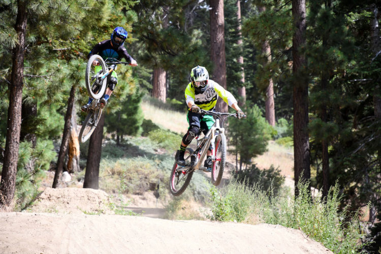 Two mountain bikers performing jumps on a dirt course surrounded by tall trees. One rider is airborne, showcasing a trick, while the other is in mid-jump, both wearing protective gear and vibrant clothing. The scene captures the excitement of outdoor biking in a forested environment.