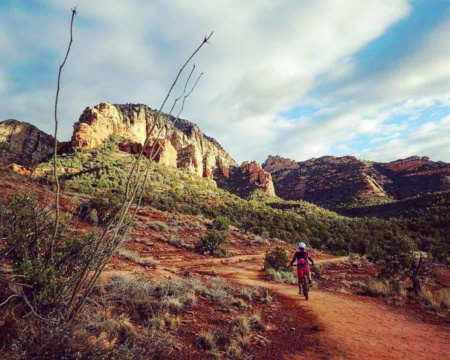 A cyclist riding on a dirt trail surrounded by rocky cliffs and lush greenery under a partly cloudy sky. The landscape features vibrant red soil and unique rock formations, creating a picturesque outdoor scene. Chuck Wagon mountain bike trail.