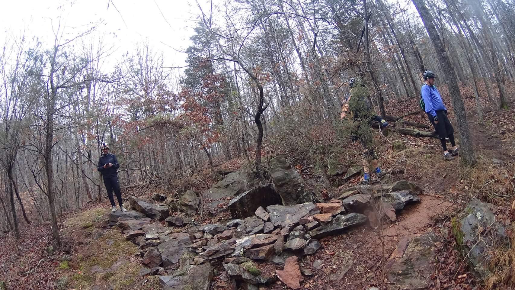 A winding trail surrounded by trees and rocks, with a few mountain bikers standing on the path, preparing to navigate the terrain. The season appears to be autumn or early winter, with leaves scattered on the ground. Mountain Laurel Trails mountain bike trail.
