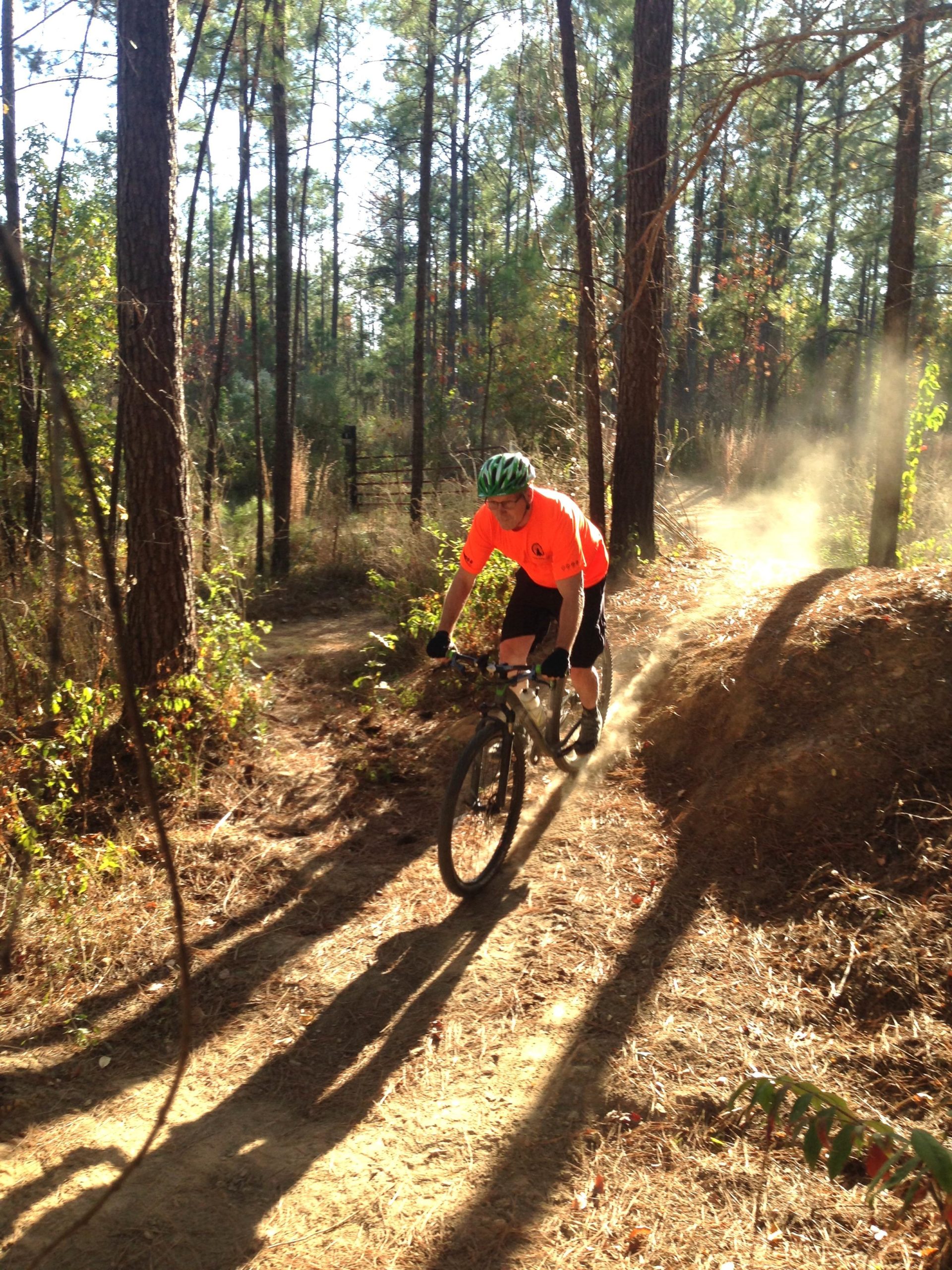 A mountain biker in an orange shirt and green helmet navigates a dirt trail through a forest, sending up dust in the sunlight filtering through the trees. Mt. Zion Bike Trails mountain bike trail.
