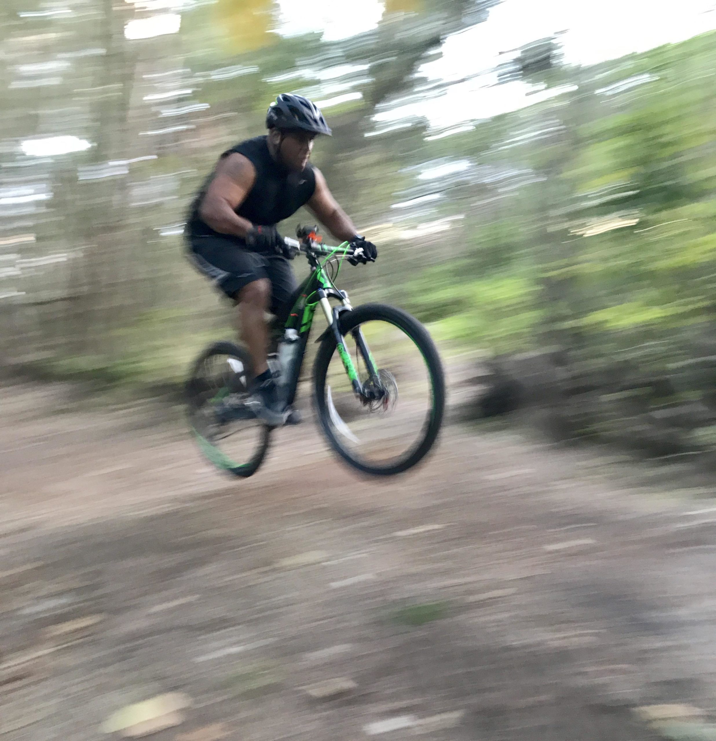 A person riding a mountain bike performs a jump on a dirt trail surrounded by trees, captured in motion with a blurred background to emphasize speed. Amelia Earhart Park mountain bike trail.